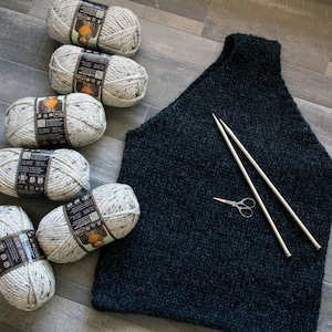 May include: A dark gray sleeveless knitted sweater with a high neck, two knitting needles, and a pair of scissors. Six balls of white yarn with brown flecks are in the background.