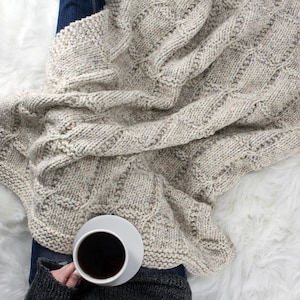 以下が含まれることがあります： ケーブル編みのパターンが付いたベージュのニットブランケットに包まれた人のクローズアップ。その人は、黒いコーヒーが入った白いマグカップを持っています。