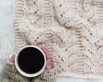 May include: A white knitted blanket with a textured pattern, a white mug with black coffee is being held by a person wearing a knitted sweater.