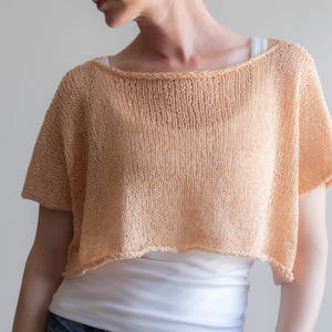 Pode incluir: Um top curto de malha cor de pêssego com mangas curtas e decote largo. O top curto é feito de um tecido leve de trama aberta. A modelo está vestindo um top branco e jeans azul.