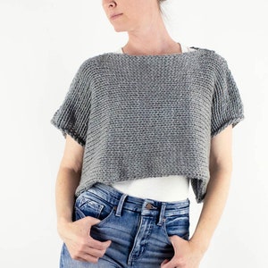 May include: A gray cropped sweater with short sleeves and a relaxed fit. The sweater is worn over a white tank top and blue jeans.