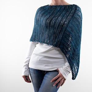 May include: A teal blue knitted poncho with a loose, open weave. The poncho is draped over a white long-sleeved shirt and blue jeans. The knitted design features a textured, ribbed pattern. The garment is lightweight and appears to be handmade.