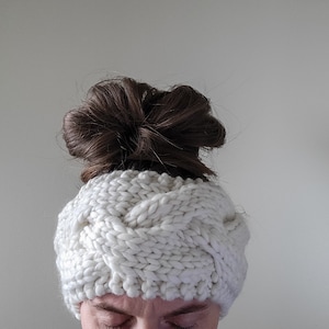 May include: A white knitted headband with a braided design. The headband is worn by a person with brown hair pulled back in a bun.