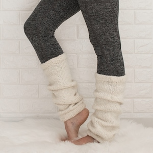 May include: A pair of white knitted leg warmers worn over grey leggings. The leg warmers are folded down at the top and bottom, creating a gathered look.