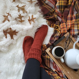 May include: A pair of cosy, rust-coloured knitted socks with a cable knit pattern. The socks are worn by a person sitting on a white fluffy rug with a plaid blanket and a cup of coffee on a wooden tray.