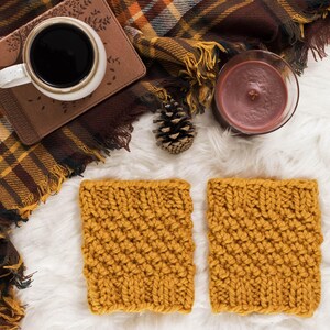 May include: Overhead shot of a cozy autumn scene. Two mustard yellow knitted coasters sit on a white faux fur surface. A mug of coffee, a candle, a pinecone, and a plaid blanket complete the warm aesthetic. The scene evokes a sense of comfort and relaxation.