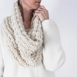 May include: A cream-colored knitted infinity scarf made of soft yarn. The scarf is worn by a person, but the person's face is not visible.