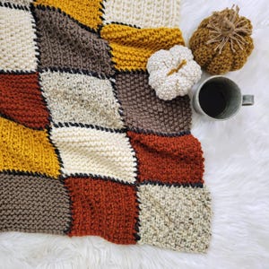 May include: A knitted patchwork blanket with squares in shades of brown, beige, yellow, red, and orange. The blanket is on a white fluffy surface with two small knitted pumpkins and a cup of coffee.