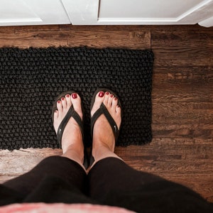 Peut inclure: Vue de dessus d'un tapis noir texturé sur un parquet. Une personne porte des tongs noires avec un logo Nike blanc. Les pieds de la personne sont visibles, avec du vernis à ongles rouge sur les orteils. Un meuble blanc est en arrière-plan.