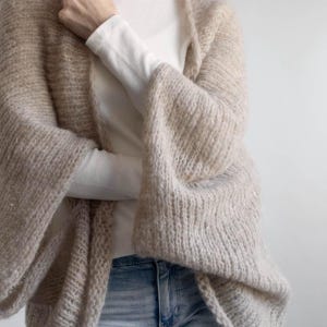 May include: A beige, open-front cardigan with wide sleeves. The cardigan is made of a soft, textured knit material. The sleeves are long and flared. The cardigan is worn over a white long-sleeved shirt and blue jeans.