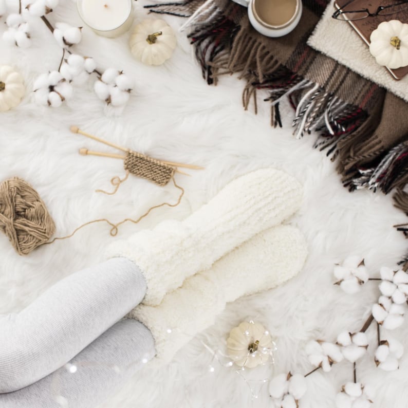 May include: White fluffy socks with a pair of knitting needles and yarn on a white fluffy surface. There are also white cotton balls and a small white pumpkin.