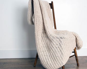 May include: A cream-colored crocheted throw blanket draped over a wooden chair. The blanket is made with a chunky yarn and has a simple, textured stitch pattern.