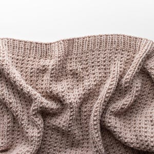 May include: A close-up shot of a knitted blanket in a neutral, light brown color. The blanket has a textured pattern with visible stitches and a ribbed border. The fabric is soft and appears to be made of wool or a similar material.