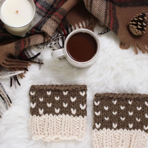 May include: Overhead shot of a cozy scene featuring two brown and cream-colored knitted cup sleeves with white heart-shaped designs. A mug of hot chocolate, a lit candle, and a plaid scarf complete the warm, inviting aesthetic.