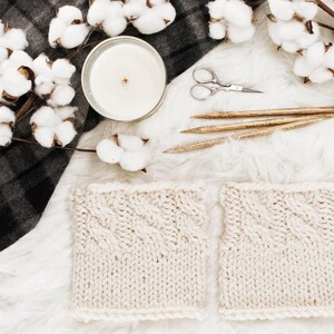 May include: A flat lay featuring a lit candle, cotton stems, knitting needles, and small scissors. Two cream-coloured knitted squares with a cable knit pattern are in the foreground. A black and grey plaid blanket is partially visible.