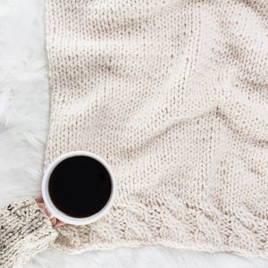 May include: A white ceramic mug filled with black coffee sits on a white knitted blanket.