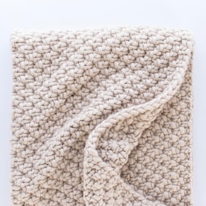 May include: A cream-colored knitted throw blanket with a textured weave. The blanket is folded in half and the edges are visible.