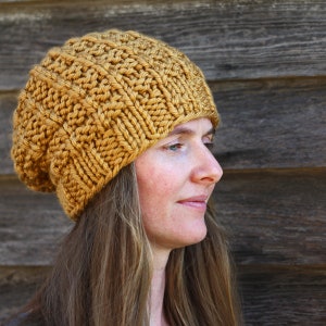 Może przedstawiać: Musztardowa dzianinowa czapka typu beanie. Czapka wykonana jest z grubej przędzy o teksturowanym, żebrowanym wzorze. Czapka jest noszona przez osobę z długimi, jasnobrązowymi włosami. Tłem jest drewniana ściana.