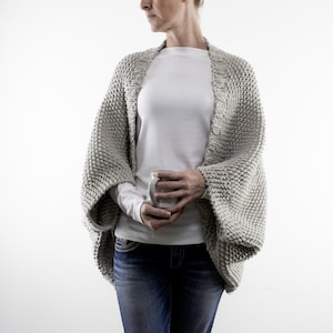 May include: A light grey, chunky knit shawl with wide sleeves. The shawl is worn over a white long-sleeved top and blue jeans. The shawl appears to be hand-knitted with a thick yarn, creating a textured appearance. The overall style is casual and comfortable.