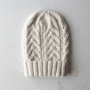 Puede incluir: Un gorro de punto de color crema con un patrón de punto de cable.