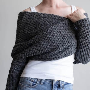 Puede incluir: Un bolero de punto gris carbón oscuro con un patrón de textura tipo gofre en el cuerpo principal y mangas acanaladas. La prenda se lleva sobre una camiseta de tirantes blanca lisa y vaqueros azules, envolviendo la parte superior del torso y los hombros.