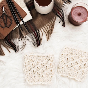May include: An overhead shot features two cream-colored, textured, knitted coasters. A brown leather journal, scissors, and knitting needles rest on a plaid blanket. A white mug and a dark red candle are also visible.