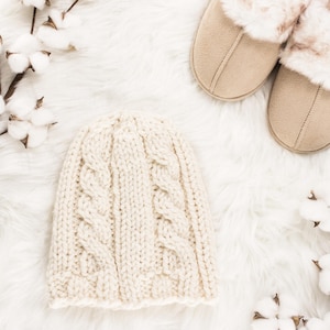 May include: A white knitted beanie with a cable knit pattern, a pair of beige suede slippers with faux fur trim, and cotton bolls on a white fluffy background.