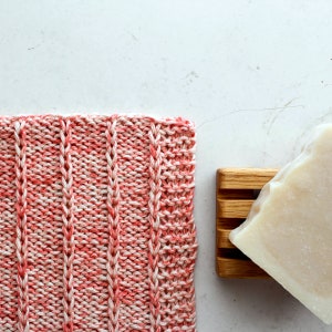 Op de afbeelding: Een roze en witte gebreide vaatdoek met een textuurpatroon. De vaatdoek ligt op een wit oppervlak naast een stuk zeep.