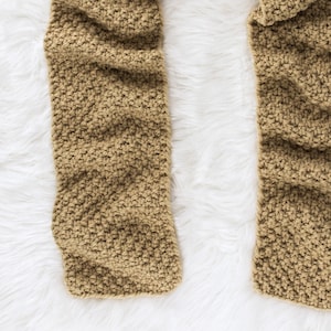 May include: A brown knitted scarf with a textured pattern. The scarf is laid out on a white fluffy surface.