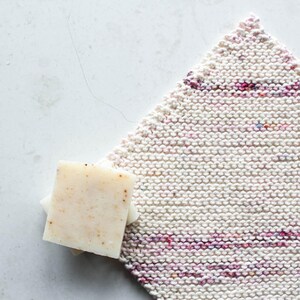 Op de afbeelding: Twee witte zeepstaven met een gespikkelde textuur, liggend op een witte gebreide theedoek met roze en paarse strepen.