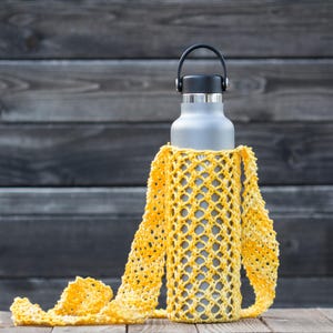 May include: A grey water bottle with a black handle and lid is in a yellow crocheted bottle holder. The holder has a strap and a net-like pattern. The bottle is on a wooden surface. The holder is made of a yellow and white yarn.