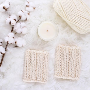 May include: A top-down view shows two cream-coloured knitted cozies, a lit candle in a glass jar, a ball of yarn, and cotton branches on a white, fluffy surface. The cozies are rectangular with a ribbed knit design, suggesting a cosy, handmade theme.