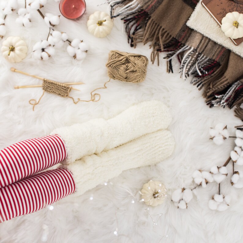 May include: White fluffy socks with red and white striped leggings on a white fluffy surface with a ball of yarn, a small knitted scarf, and cotton balls.