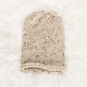 May include: A beige knitted beanie with a flecked texture. The beanie has a folded brim.
