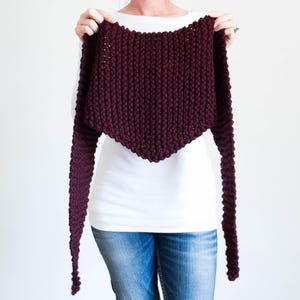 Può includere: Uno scialle lavorato a maglia bordeaux con maniche lunghe e affusolate. Lo scialle è presentato su uno sfondo bianco, evidenziando il suo design a maglia grossa e strutturata. L'indumento ha uno scollo rotondo e un bordo inferiore triangolare.