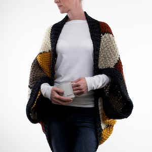 Peut inclure: Une femme portant un cardigan crocheté multicolore avec une chemise blanche à manches longues et un jean bleu. Elle tient une tasse blanche sur laquelle est inscrit "GRATEFUL".