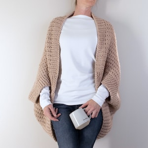 May include: A woman wearing a light brown knitted cardigan with long sleeves and a relaxed fit. She is wearing a white t-shirt and blue jeans. She is holding a white mug with the word "GRATEFUL" printed on it.
