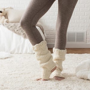 May include: A pair of white knitted leg warmers with a slouchy fit. The leg warmers are worn over brown leggings.