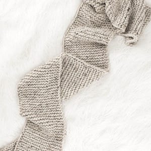 May include: A beige knitted scarf with a textured pattern, folded and displayed on a white fluffy surface.