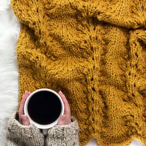 May include: A white ceramic mug filled with black coffee, held by hands wearing a beige knitted sweater. The mug is resting on a mustard yellow knitted blanket.