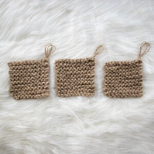 May include: Three square, hand-knitted jute pot holders with hanging loops. The pot holders are a natural brown color and are arranged on a white, fluffy surface. The knitted texture is clearly visible, showcasing the craftsmanship.