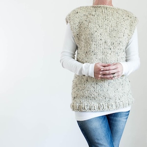 May include: A sleeveless knitted vest in a cream and brown speckled yarn. The vest has a relaxed fit and a slightly rounded neckline.