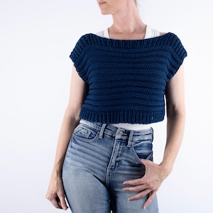 May include: A navy blue, cropped knit top with a wide neckline and short sleeves. The top is made of a textured yarn and has a ribbed hem. The model is wearing blue jeans.