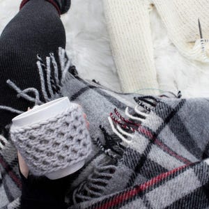 May include: A white ceramic mug with a grey knitted cosy. The mug is being held by a person wrapped in a grey and black plaid blanket with red accents.