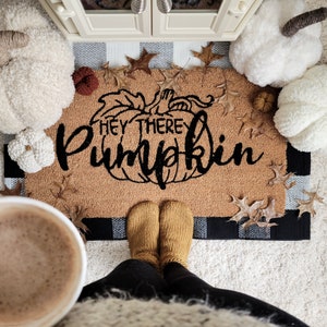 May include: A brown doormat with the text "They There Pumpkin" in black lettering. The doormat has a black and white checkered border. A pair of brown fuzzy slippers are on the doormat.