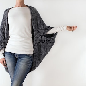 May include: A grey knitted cardigan with long sleeves and a wide, open front. The cardigan is worn over a white t-shirt and blue jeans.