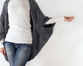 May include: A gray knitted cardigan with long sleeves and a wide, open front. The cardigan is worn over a white t-shirt and blue jeans.