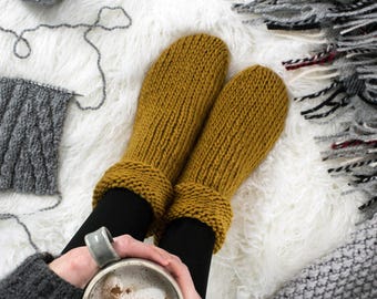 May include: A pair of cozy, mustard yellow knitted slippers with a ribbed texture. The slippers are worn by a person holding a mug of hot chocolate with marshmallows.
