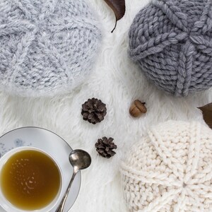 May include: A top-down view shows three knitted hats in shades of grey and cream, set on a white, fluffy background. A cup of tea with a spoon, pine cones, and an acorn add to the autumnal scene. The hats have a textured cable-knit pattern.
