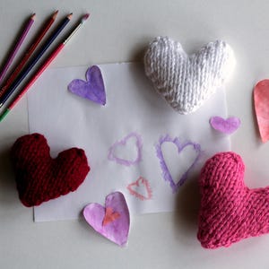 May include: A white sheet of paper with watercolour hearts in shades of pink, purple, and white. Three knitted hearts are placed on the paper, one red, one white, and one pink.